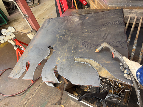 A plasma cutter nozzle is held in a gloved hand in front of the sheet. Four tentacles have been cut out